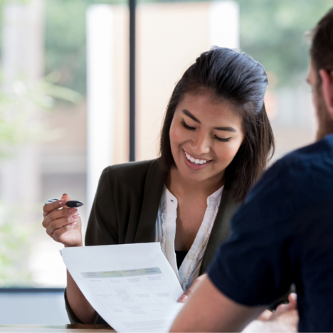 A professional woman in a business setting is smiling while reviewing documents with a colleague across the table.