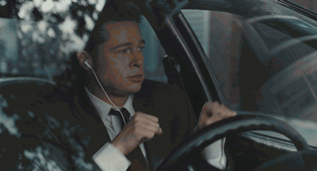 A man in a suit and tie dances and gestures energetically while sitting inside a car, listening to music through earbuds, with buildings visible outside the windows.