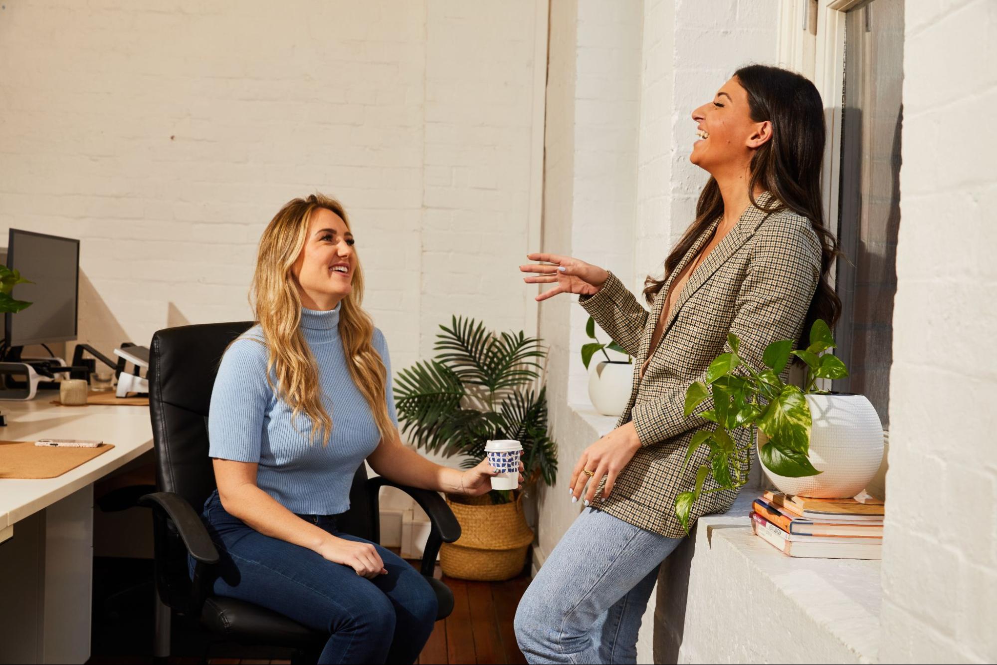 Two women converse; one sits holding a cup, smiling, while the other leans on a window ledge, gesturing. The bright office features potted plants, books, and computer desks.