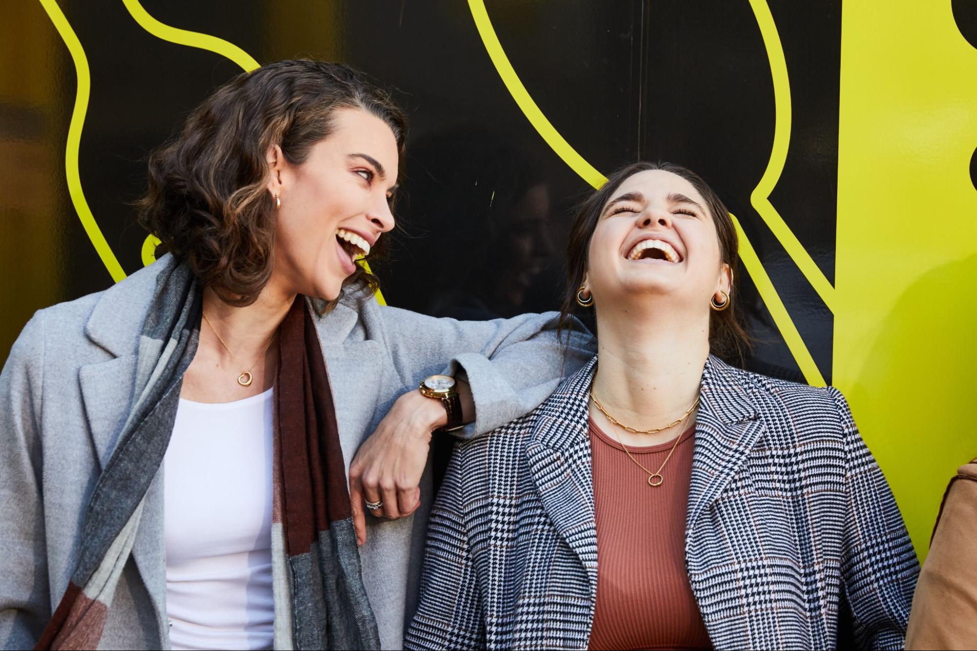 Two women are standing side by side, both wearing coats. The first woman has her elbows resting on the other woman's shoulder and is looking at her while laughing. The second woman is laughing with her head tilted back and her eyes closed. Behind them is a black wall covered with yellow lines and patterns.
