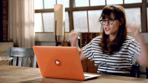 A woman with glasses is enthusiastically raising her arms in front of an orange laptop with a play button logo, sitting at a wooden table in a room with large windows.